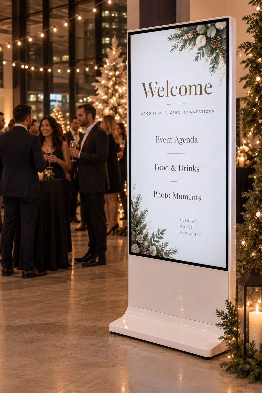 Tall white vertical display at a corporate holiday party, guests mingling near string lights and holiday greenery, screen showing a welcome sign with event agenda, food and drinks, and photo moments listed
